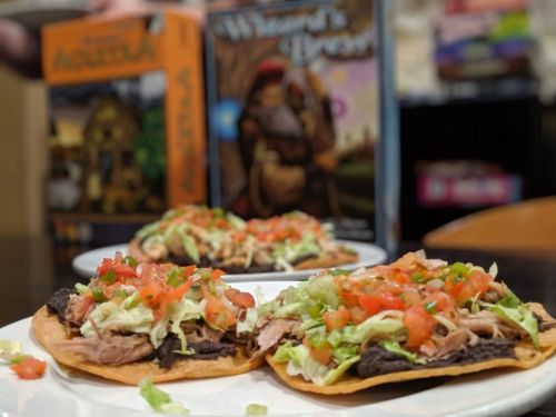 Two tacos topped with shredded chicken, lettuce, and diced tomatoes, with board games in the background.