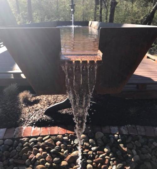 A modern water feature with a rectangular basin, flowing water, and pebbled ground, surrounded by greenery.