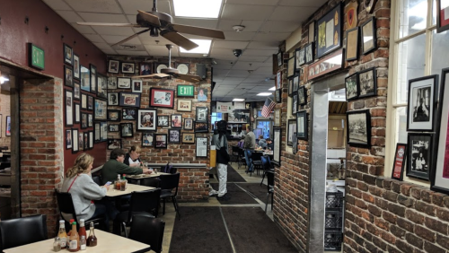 A cozy restaurant interior with brick walls, framed photos, and diners at tables. A fan hangs from the ceiling.