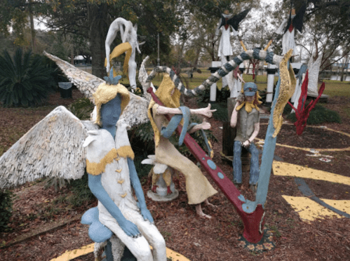 Colorful sculptures of figures with wings and unique features in a park setting, surrounded by trees and pathways.