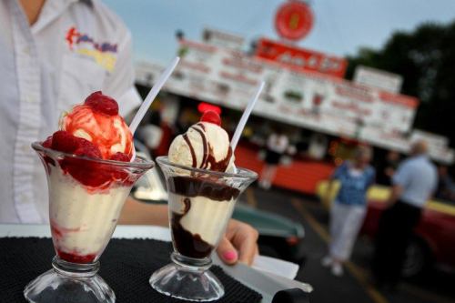 Two ice cream sundaes in glass cups, one topped with raspberries and the other with chocolate sauce, served at a food stand.