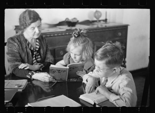 A woman and two children sit at a table, reading a book together in a well-lit room.