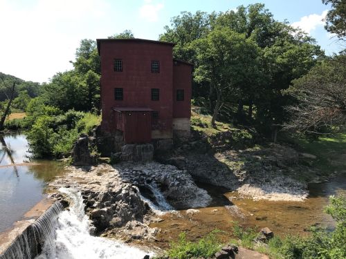 A red, historic building beside a small waterfall and stream, surrounded by lush green trees and a clear blue sky.