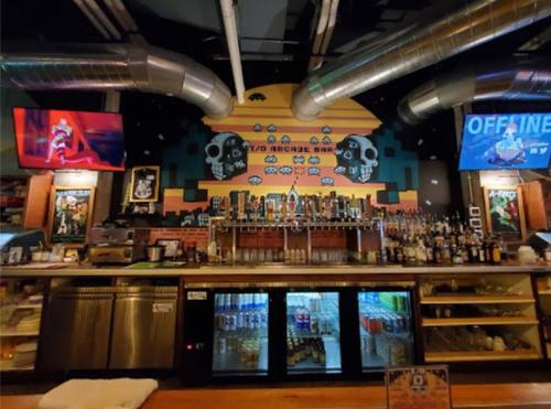A vibrant bar with a retro arcade theme, featuring colorful wall art, multiple screens, and a well-stocked drink counter.