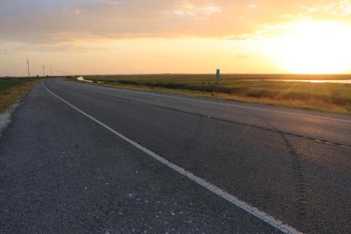 A winding road stretches into the distance under a vibrant sunset, with grassy fields and water on either side.