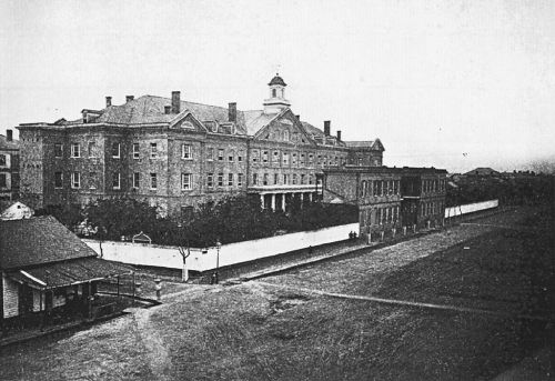 Historic black-and-white photo of a large building with a cupola, surrounded by trees and a white fence.