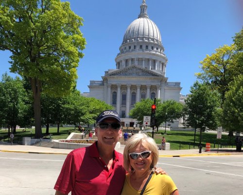A couple poses in front of a large, domed government building on a sunny day, surrounded by green trees.