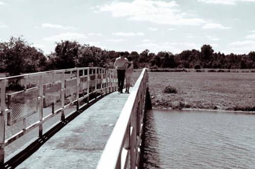 A couple walks hand in hand on a bridge over a calm body of water, surrounded by greenery and a clear sky.