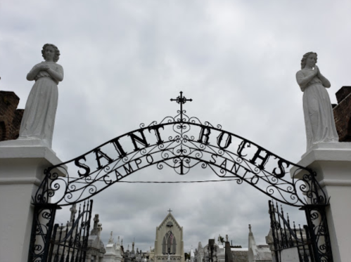 Gates of Saint Roch's Cemetery, featuring white statues and a cross, under a cloudy sky.