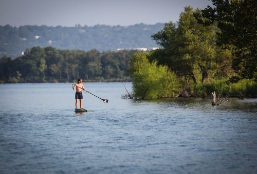 A person paddleboarding on a calm lake surrounded by trees and greenery under a clear sky.