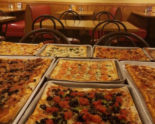 A variety of pizzas on trays, featuring different toppings, in a cozy restaurant setting with red booths.