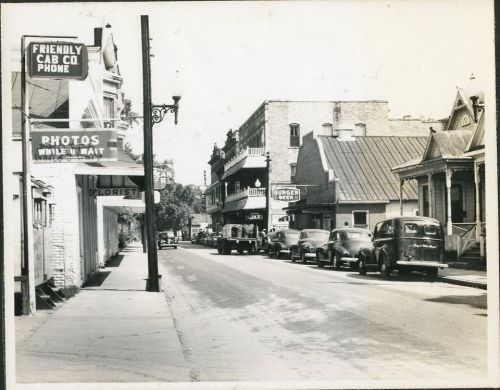 Black and white photo of a vintage street scene with parked cars, storefronts, and a cab company sign.