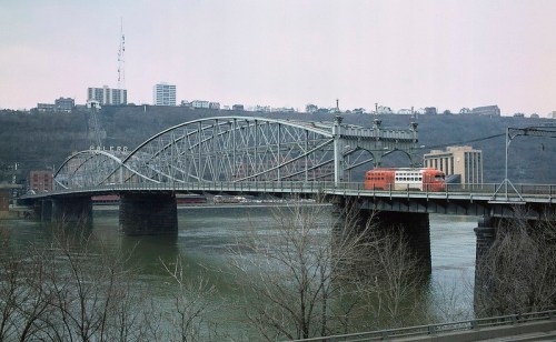 A vintage train crosses a steel arch bridge over a river, with a city skyline and trees in the background.