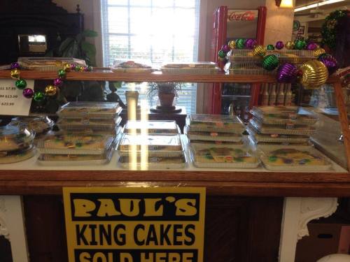 Display of king cakes in clear containers, decorated with festive beads, with a sign reading "Paul's King Cakes Sold Here."