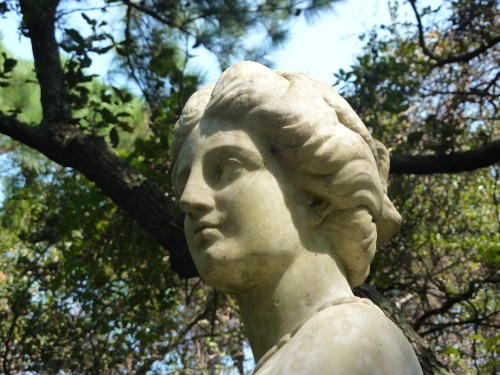 A close-up of a weathered stone statue of a woman, surrounded by greenery and soft sunlight.