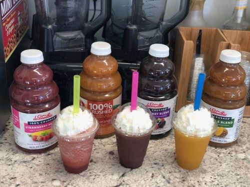 Three colorful smoothies topped with whipped cream and straws, alongside bottles of fruit juice in the background.