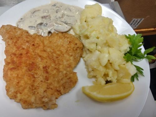 A plate featuring a breaded fish fillet, creamy mushroom sauce, mashed potatoes, and a lemon wedge, garnished with parsley.