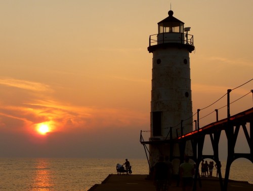 Silhouette of a lighthouse at sunset, with people walking along the pier and a calm sea in the background.