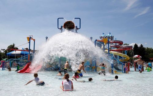 Children play in a water park, splashing in a pool with slides and a large water feature under a sunny sky.