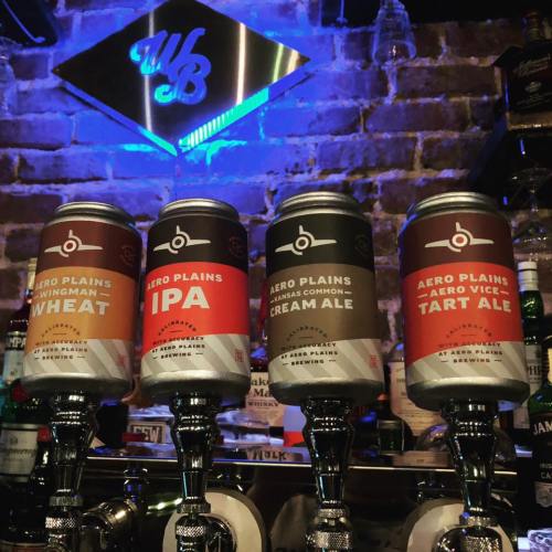 Four cans of Aero Plains beer on a bar, featuring Wingman Wheat, IPA, Cream Ale, and Tart Ale, with a blue-lit backdrop.