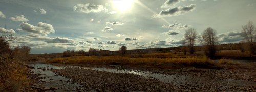 A serene landscape featuring a river, rocky shore, autumn trees, and a cloudy sky with the sun shining above.