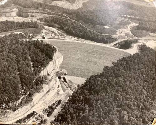 Aerial view of a landscape featuring a river, forested hills, and a large cleared area, possibly a quarry or construction site.