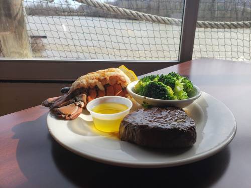 A plate featuring a lobster tail, grilled steak, broccoli, and a small cup of olive oil, with a scenic view in the background.