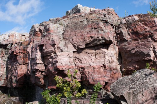 A rugged rock formation with various colors and textures, surrounded by greenery under a blue sky.