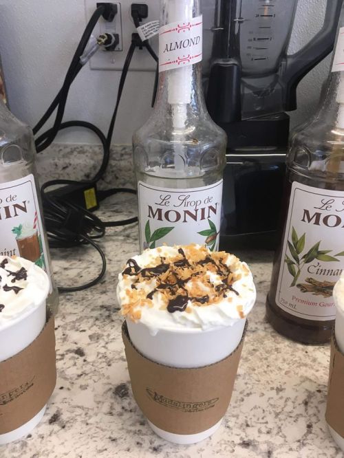 A coffee drink topped with whipped cream, chocolate drizzle, and coconut flakes, next to Monin syrup bottles.