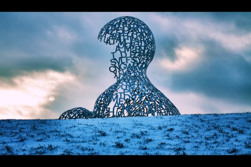 A large, abstract sculpture made of letters, set against a cloudy sky and snowy landscape.