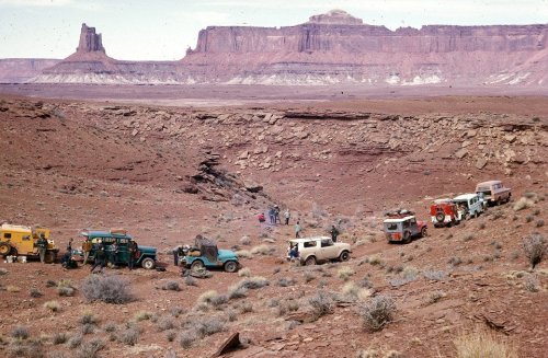 A group of vintage vehicles parked in a desert landscape with rocky formations in the background.