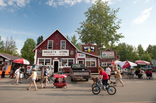 A quaint red store in Talkeetna, Alaska, with people walking, outdoor seating, and a truck parked outside.