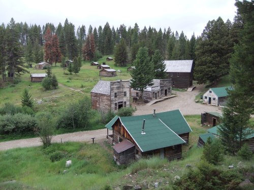 Aerial view of a rustic ghost town with wooden buildings surrounded by lush green trees and a gravel road.
