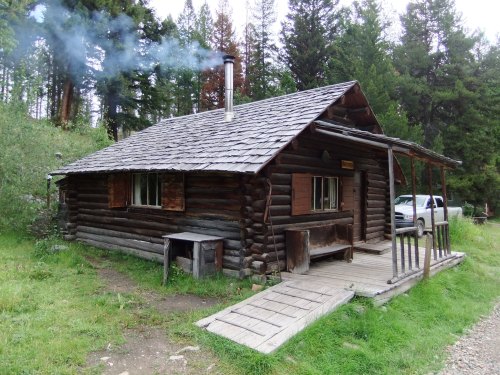 A rustic log cabin with a smoke rising from the chimney, surrounded by trees and a grassy area.