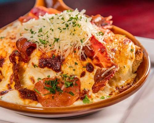 A close-up of a cheesy baked dish topped with crispy bacon, herbs, and grated cheese in a brown bowl.