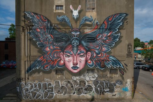 A large mural on a building featuring a woman's face with butterfly wings, an eye on her forehead, and a rabbit above.