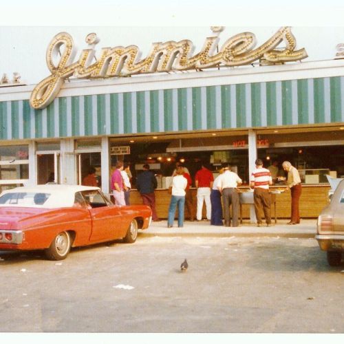 A vintage diner named "Jimmies" with a crowd outside, classic cars parked nearby, and a pigeon in the foreground.