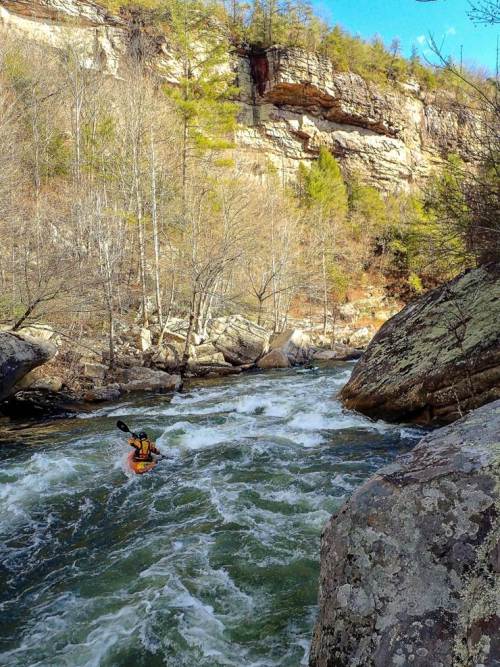 A kayaker navigates a rushing river surrounded by rocky cliffs and trees in a scenic outdoor landscape.