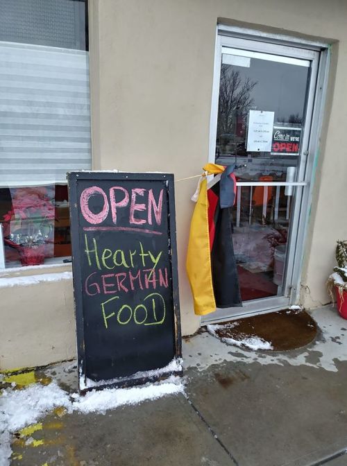 A chalkboard sign outside a restaurant reads "OPEN" and "Hearty GERMAN FOOD," with a German flag hanging nearby.