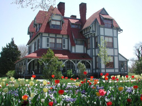 Victorian-style house with a red roof, surrounded by colorful tulips and greenery on a sunny day.