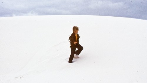 A child walks through a vast, white sand dune under a cloudy sky.