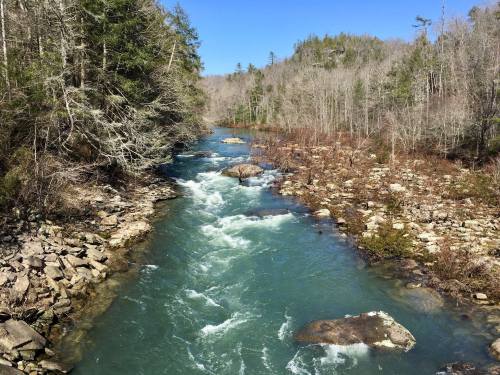 A flowing river surrounded by rocky banks and sparse trees under a clear blue sky.