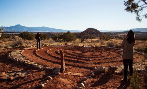 Two people stand in a stone labyrinth in a desert landscape, with mountains in the background under a clear sky.