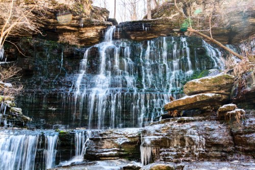 A serene waterfall cascading over rocky cliffs, surrounded by trees and ice formations in a tranquil natural setting.