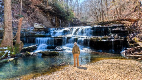 A person stands by a serene waterfall surrounded by trees, with sunlight filtering through the branches.