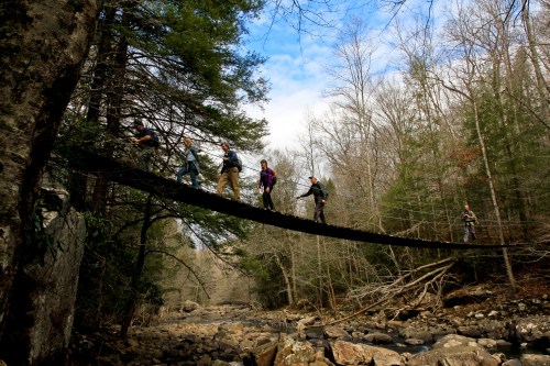 A group of hikers crosses a suspension bridge over a rocky river, surrounded by trees and a blue sky.