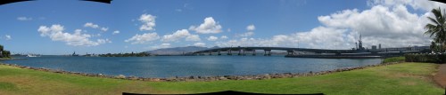 Panoramic view of a harbor with a bridge, blue water, green grass, and scattered clouds in a clear sky.