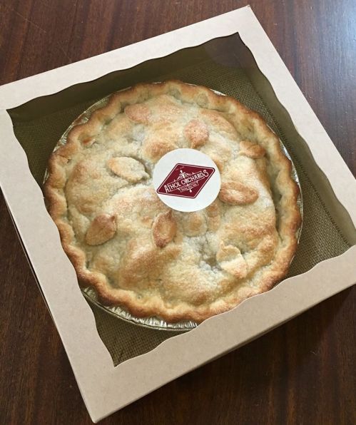 A freshly baked pie in a cardboard box with a circular label on top, resting on a wooden surface.