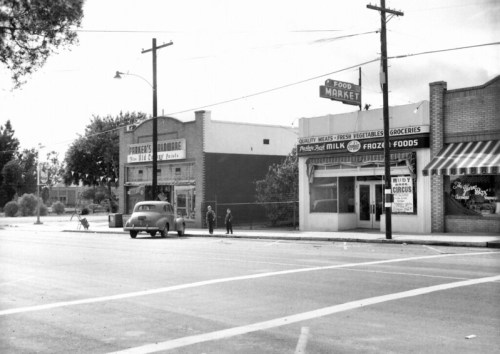 Black and white photo of a street scene featuring shops, a food market, and children playing near the road.