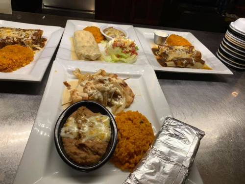 A variety of Mexican dishes on white plates, including rice, beans, enchiladas, and a salad, on a metal table.